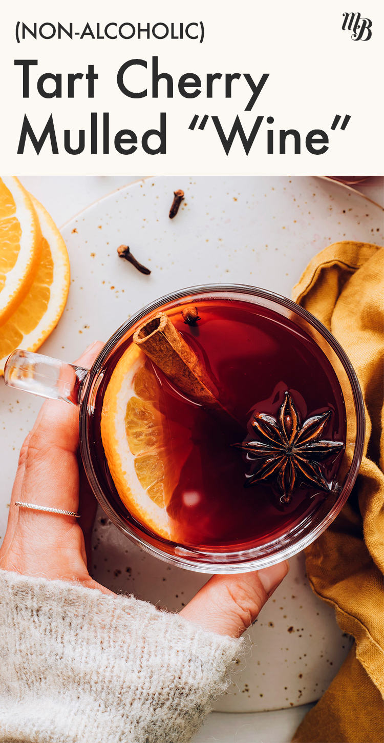 Hand holding a glass of tart cherry mulled "wine" with an orange slice, clove star, and cinnamon stick in it
