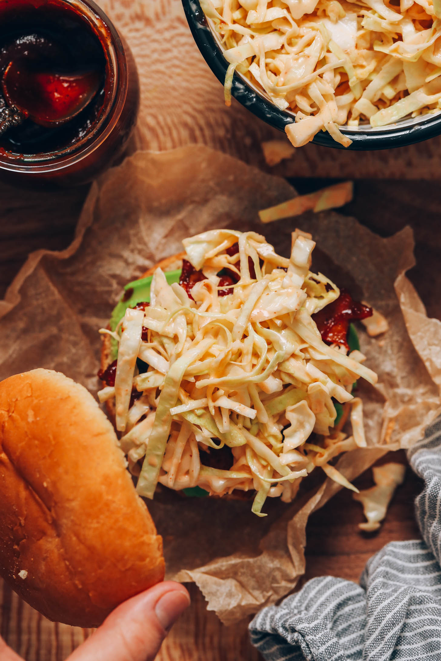 BBQ-Sauce und Krautsalat neben einer Hand, die ein Brötchen über einem Sandwich hält