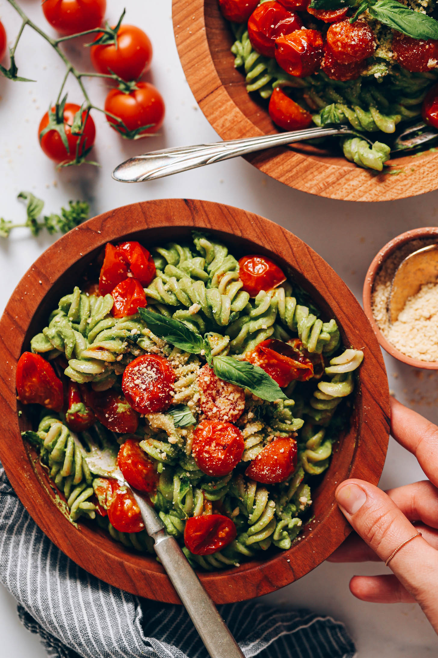 Hand resting on a bowl of creamy vegan pesto pasta with roasted tomatoes
