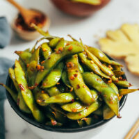 Bowl of spicy garlic edamame pods next to ginger and chili flakes