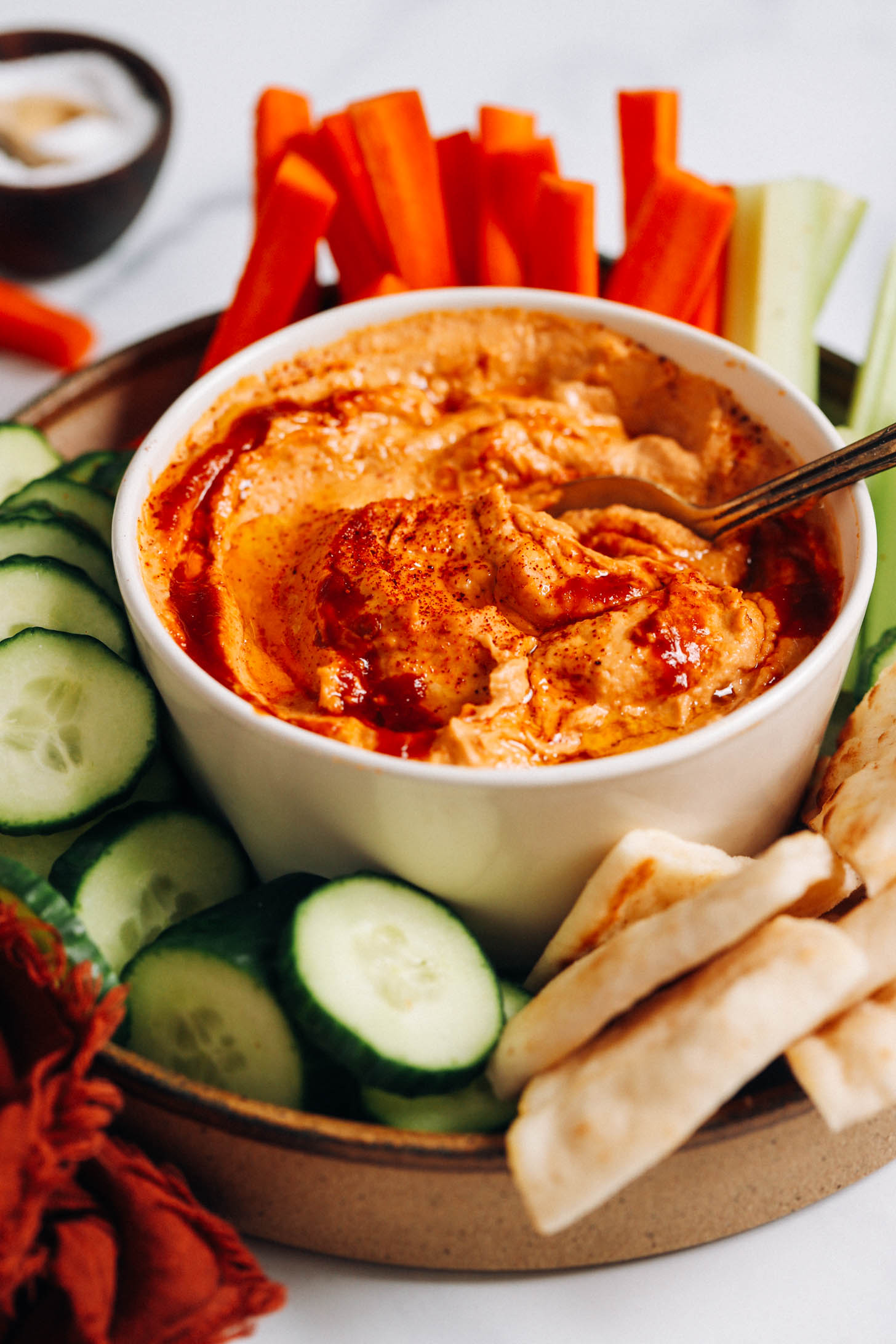 Bowl of spicy chipotle hummus surrounded by cucumber slices, carrot and celery sticks, and pita bread