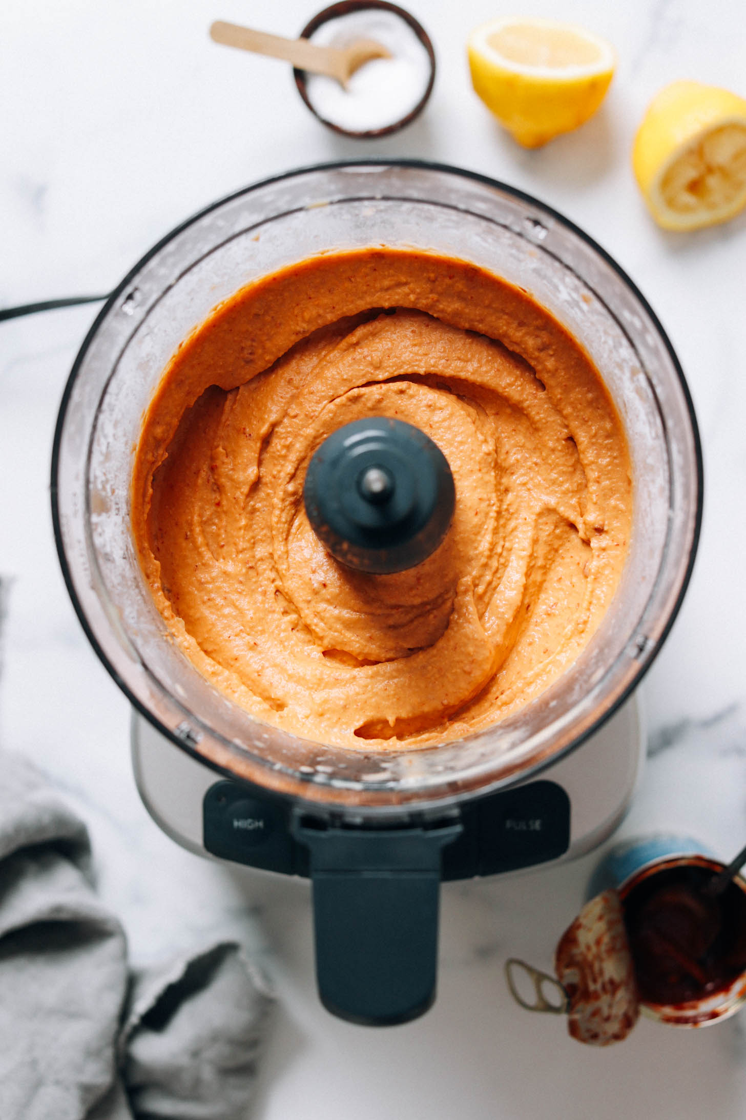 Vibrant red-orange hummus in a food processor next to lemons, salt, and adobo peppers