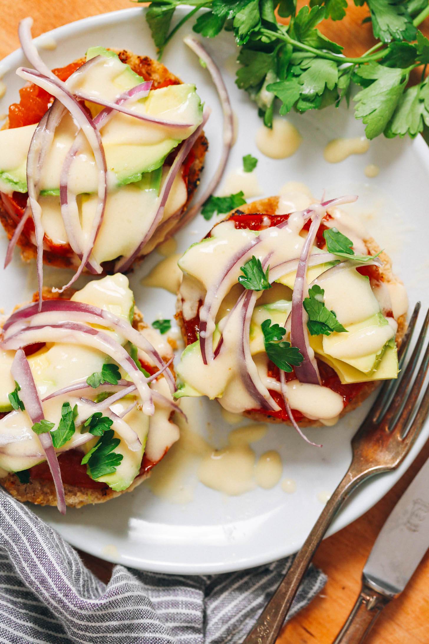 Top down shot of English muffins topped with seared tomatoes, avocado, red onion, parsley, and vegan hollandaise sauce