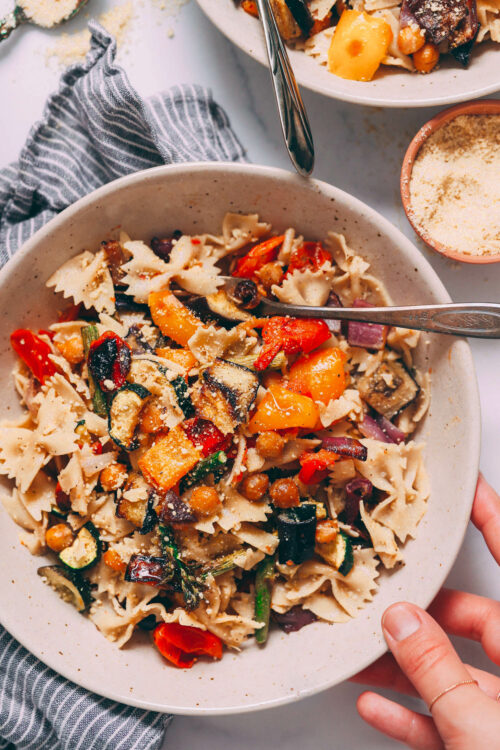 Hand holding a bowl of vegan pasta primavera with roasted veggies