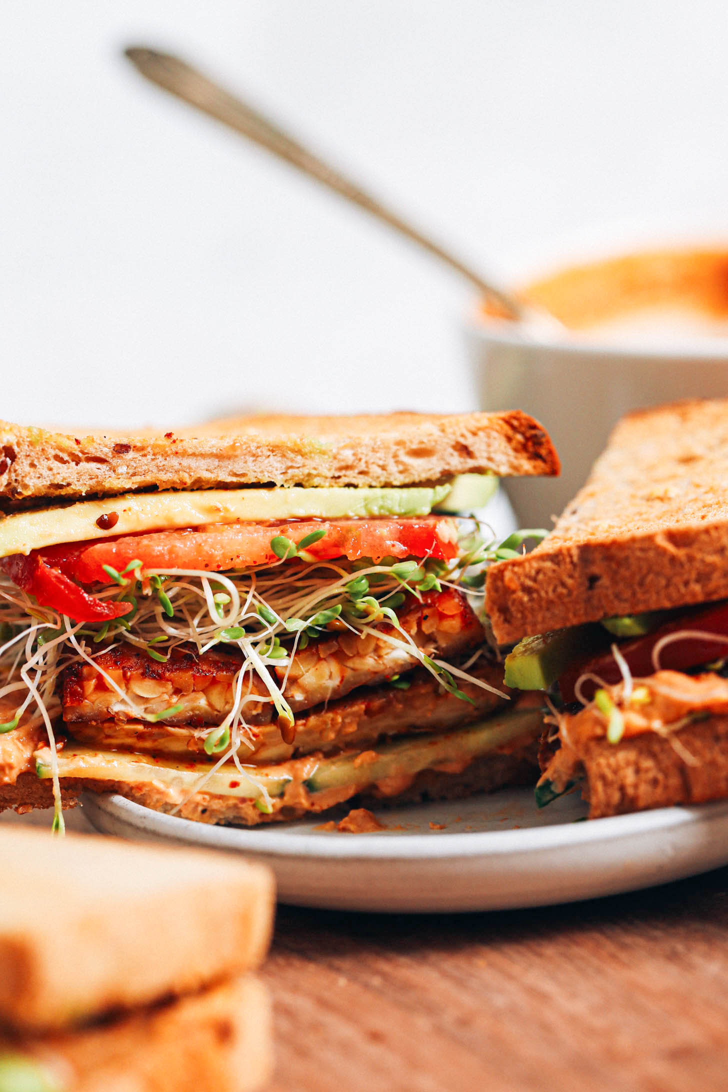 Sandwich with layers of avocado, tomato, sprouts, tempeh bacon, cucumber, and chipotle hummus