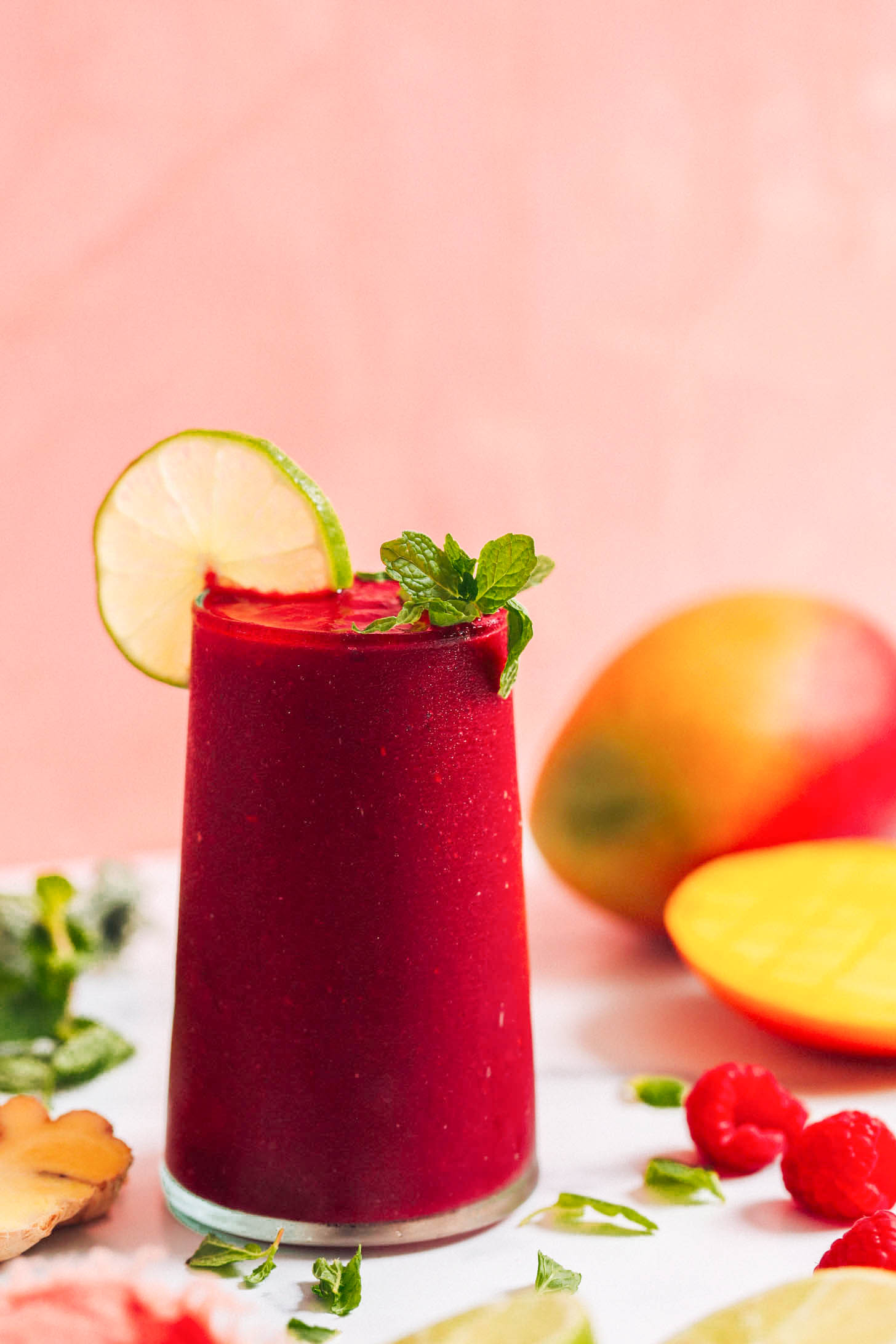 Raspberries, mango, mint, lime, and ginger surrounding a glass of our mango beet smoothie recipe