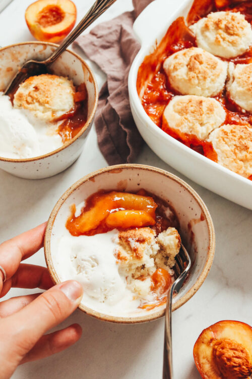 Bowl of gluten-free peach cobbler with a scoop of vegan vanilla ice cream next to a baking dish with more cobbler