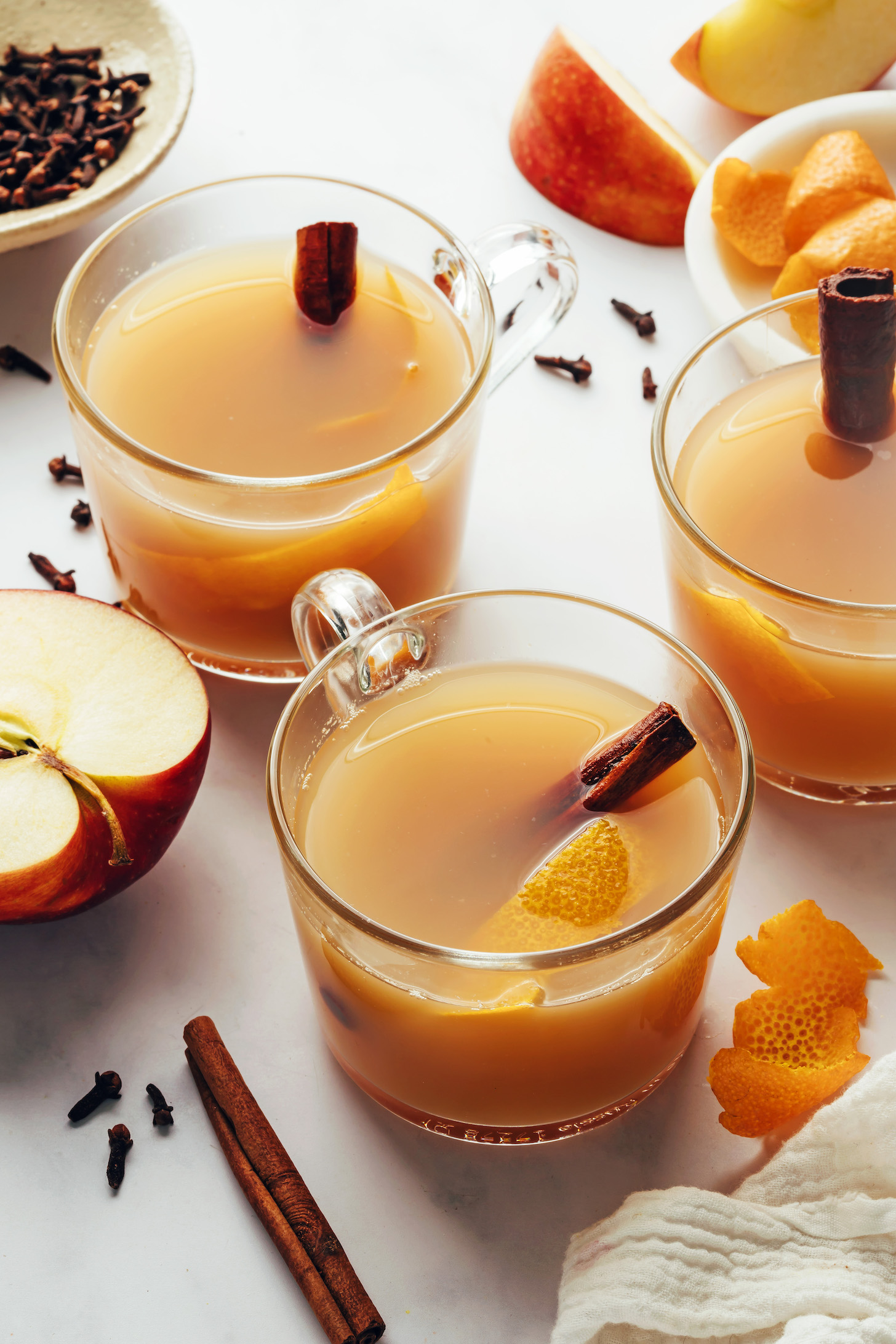 Glass mugs of hot spiced apple cider with cinnamon sticks and orange peel in and around them