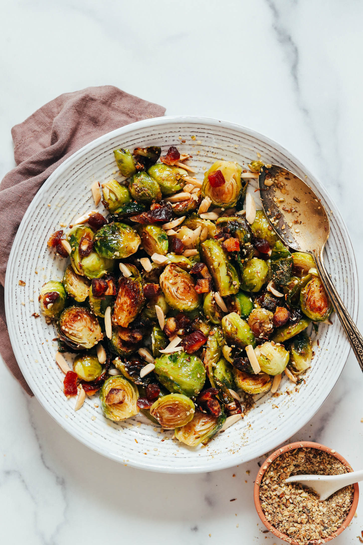 Bowl of dukkah seasoning next to a larger bowl of crispy roasted Brussels sprouts with dates and almonds