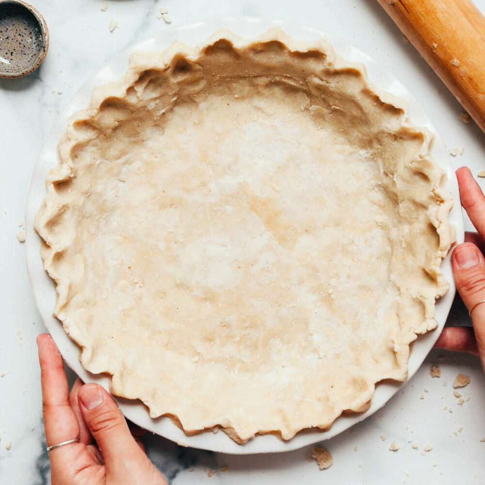 Tangan memegang bahagian tepi kuali pai yang diisi dengan kerak pai vegan dan bebas gluten buatan sendiri