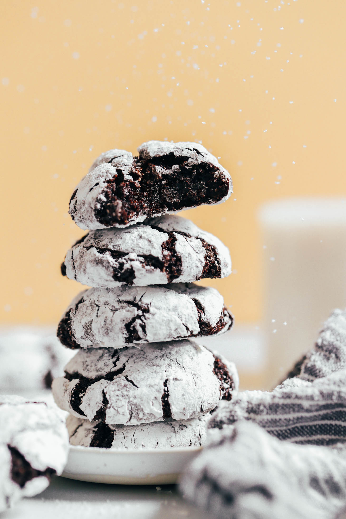Sprinkling powdered sugar on a stack of chocolate crinkle cookies with the top cookie showing the inner fudgy center