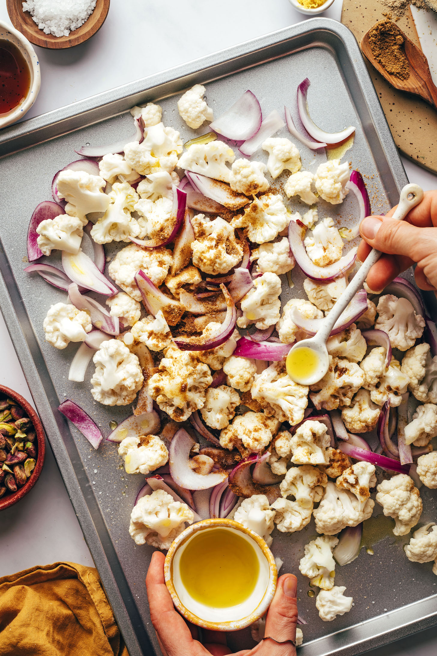 Using a spoon to add olive oil to a baking sheet of cauliflower florets, red onion, and spices