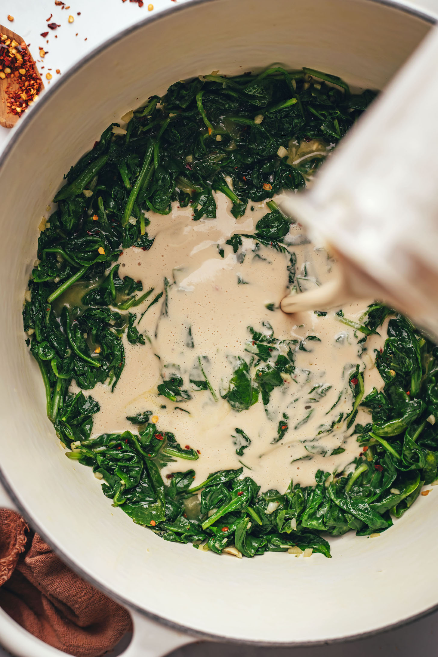Pouring a creamy cashew-based sauce over sautéed spinach