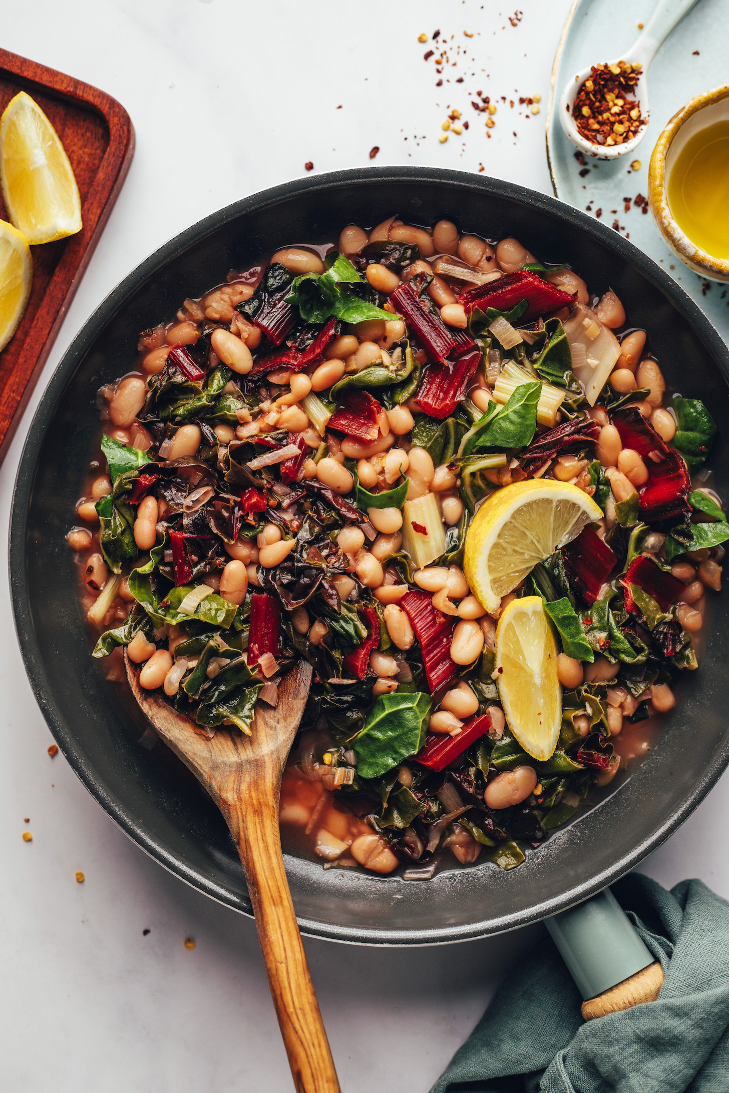 Lemons, olive oil, and red pepper flakes around a pan of beans and greens with lemon wedges