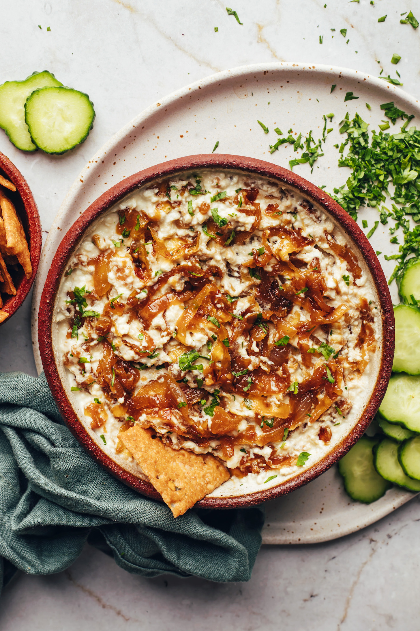 Parsley, sliced cucumbers, and crackers around a bowl of vegan caramelized onion dip