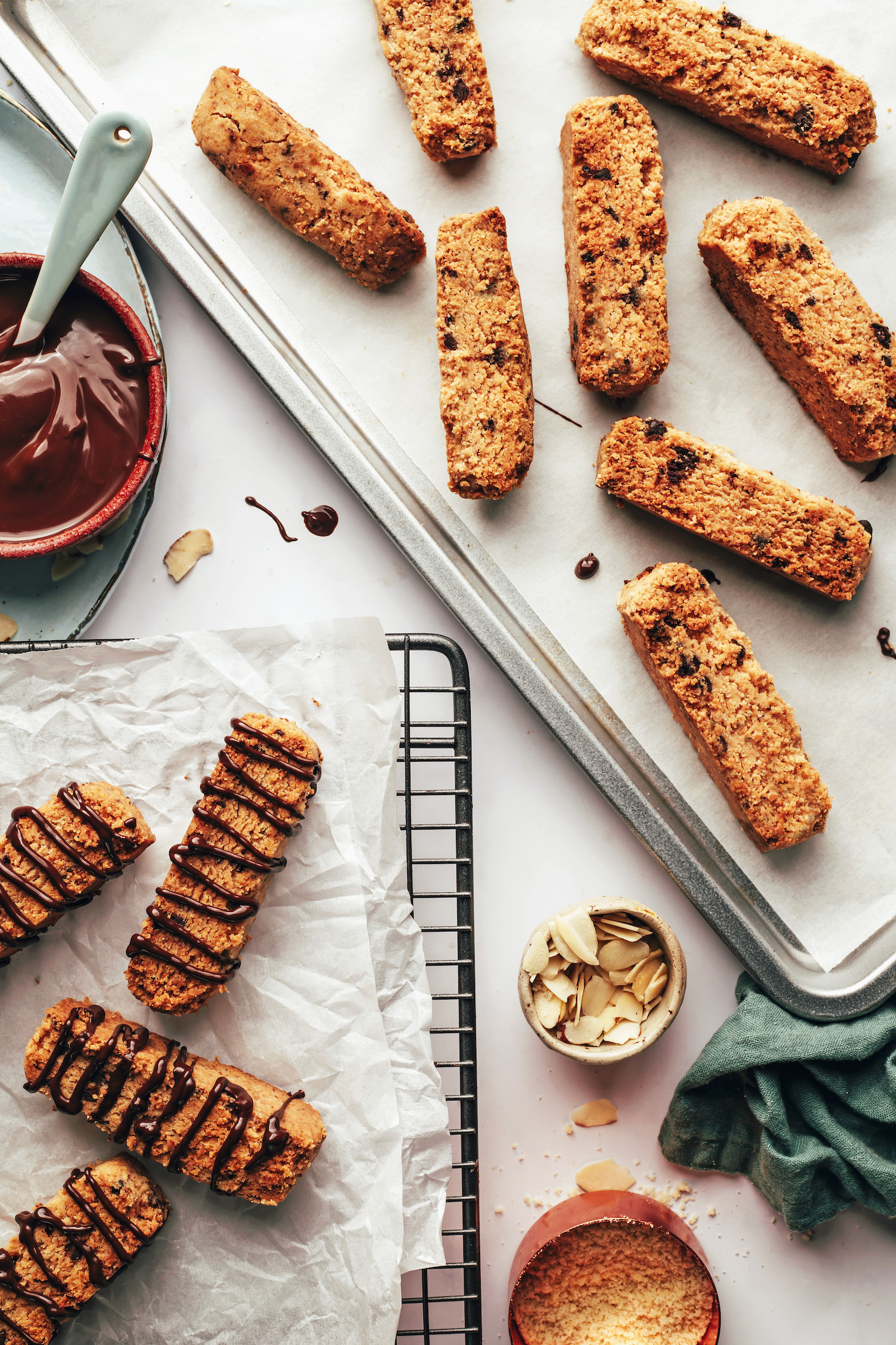 Biscotti on a parchment-lined baking sheet and on a cooling rack drizzled with chocolate