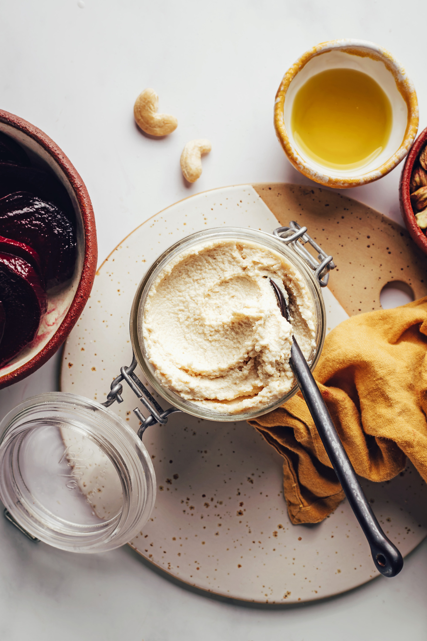 Fotografía cenital de tazones de aceite de oliva, anacardos y remolachas asadas, además de una cuchara en un frasco de queso de cabra vegano casero