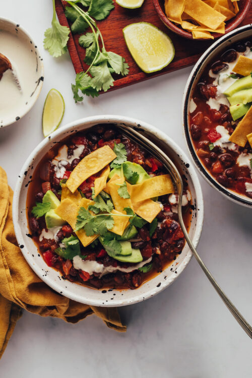 Bowls of vegan chipotle black bean chili topped with tortilla strips, cilantro, and avocado