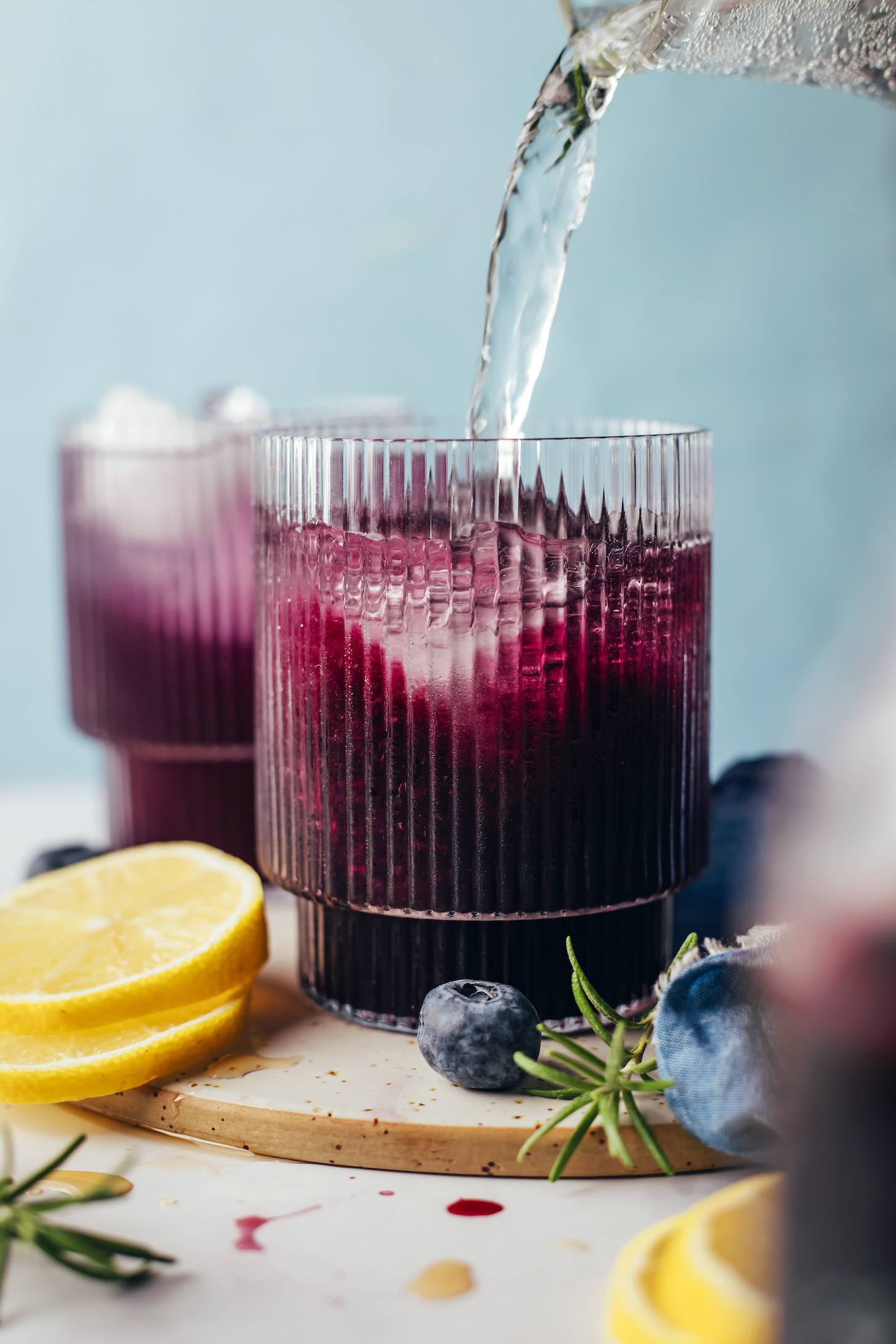 Sparkling water being poured into a glass with ice cubes and naturally sweetened blueberry simple syrup 