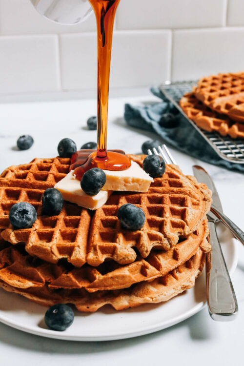 Drizzling maple syrup onto a stack of protein waffles topped with vegan butter and blueberries