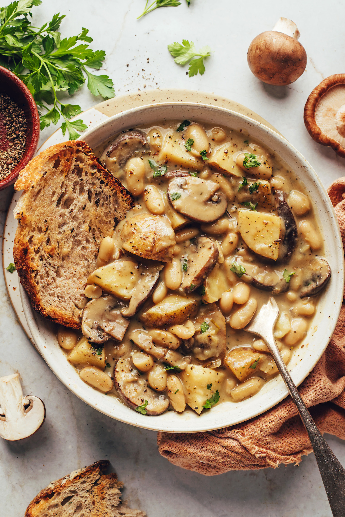 Mushrooms, parsley, and black pepper around a bowl of white bean mushroom stew with a slice of toasted bread in it