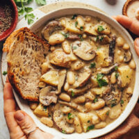 Hands holding a bowl of vegan white bean mushroom stew with a slice of toasted bread in it