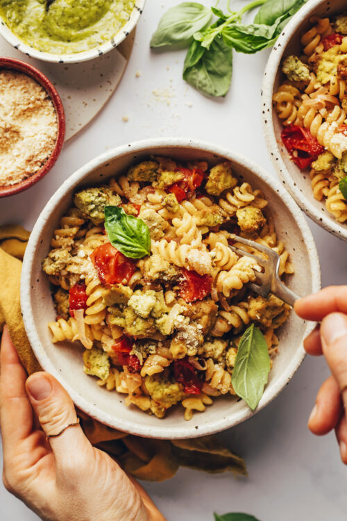 Hands holding the side of a bowl and a fork picking up a bite of our vegan tofu pesto pasta bowl