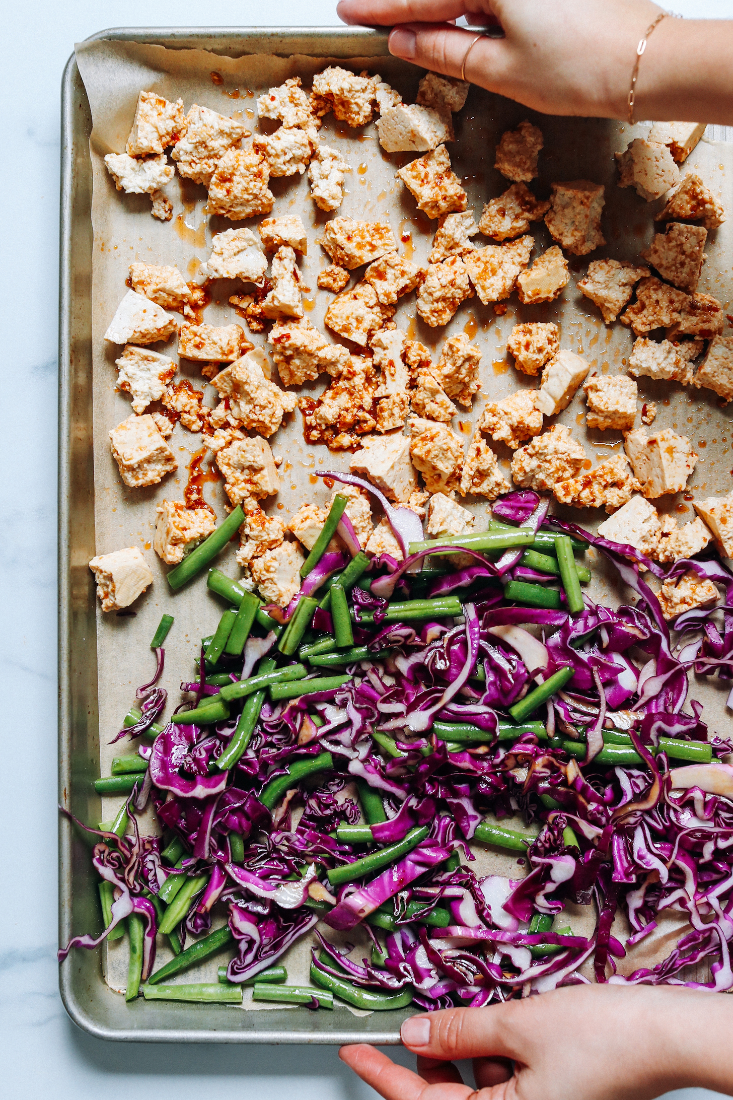 Hands holding a baking sheet of seasoned crumbled tofu, green beans, and red cabbage
