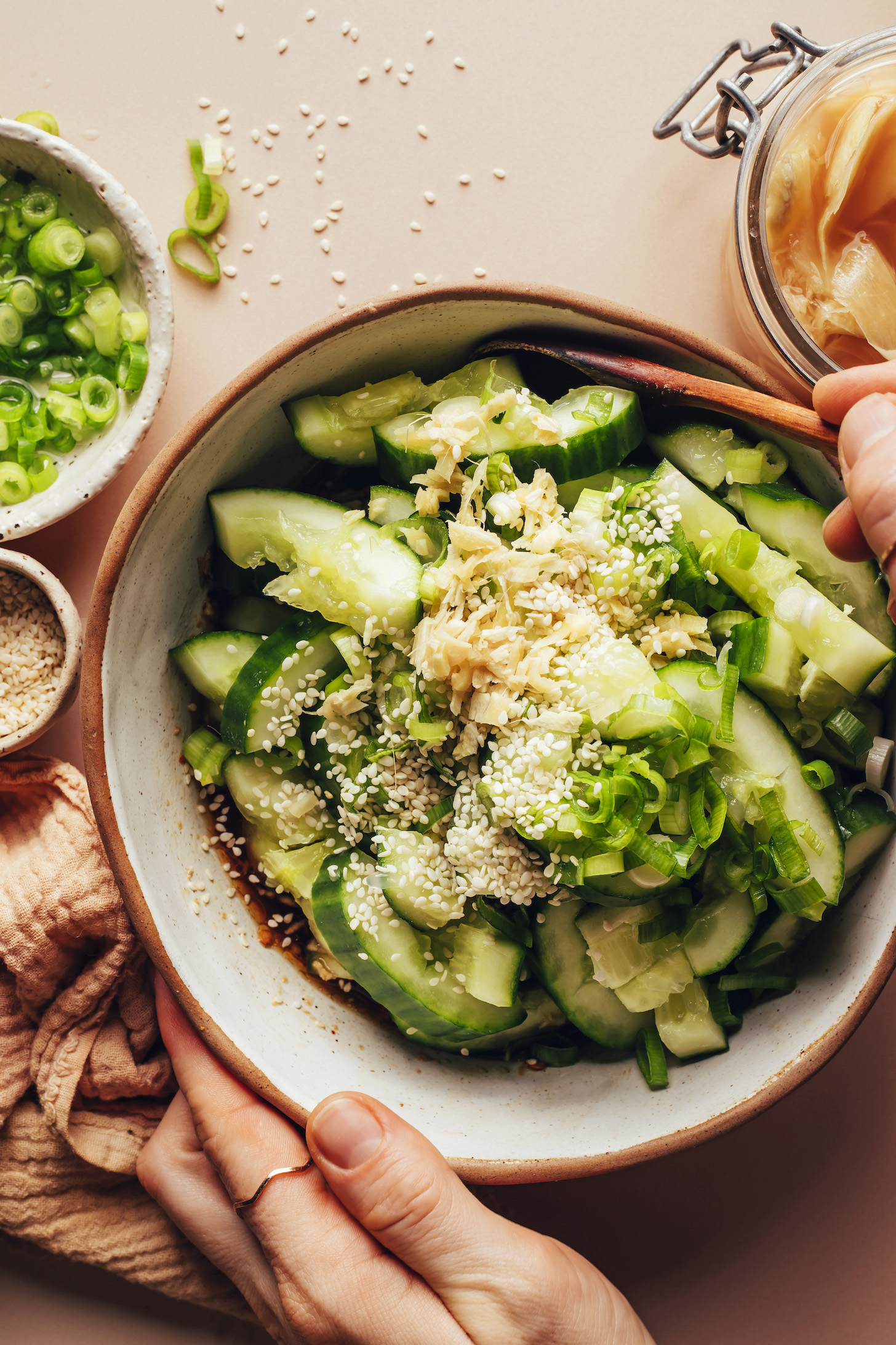Hands holding a bowl of sliced cucumbers with chopped pickled ginger and sesame seeds