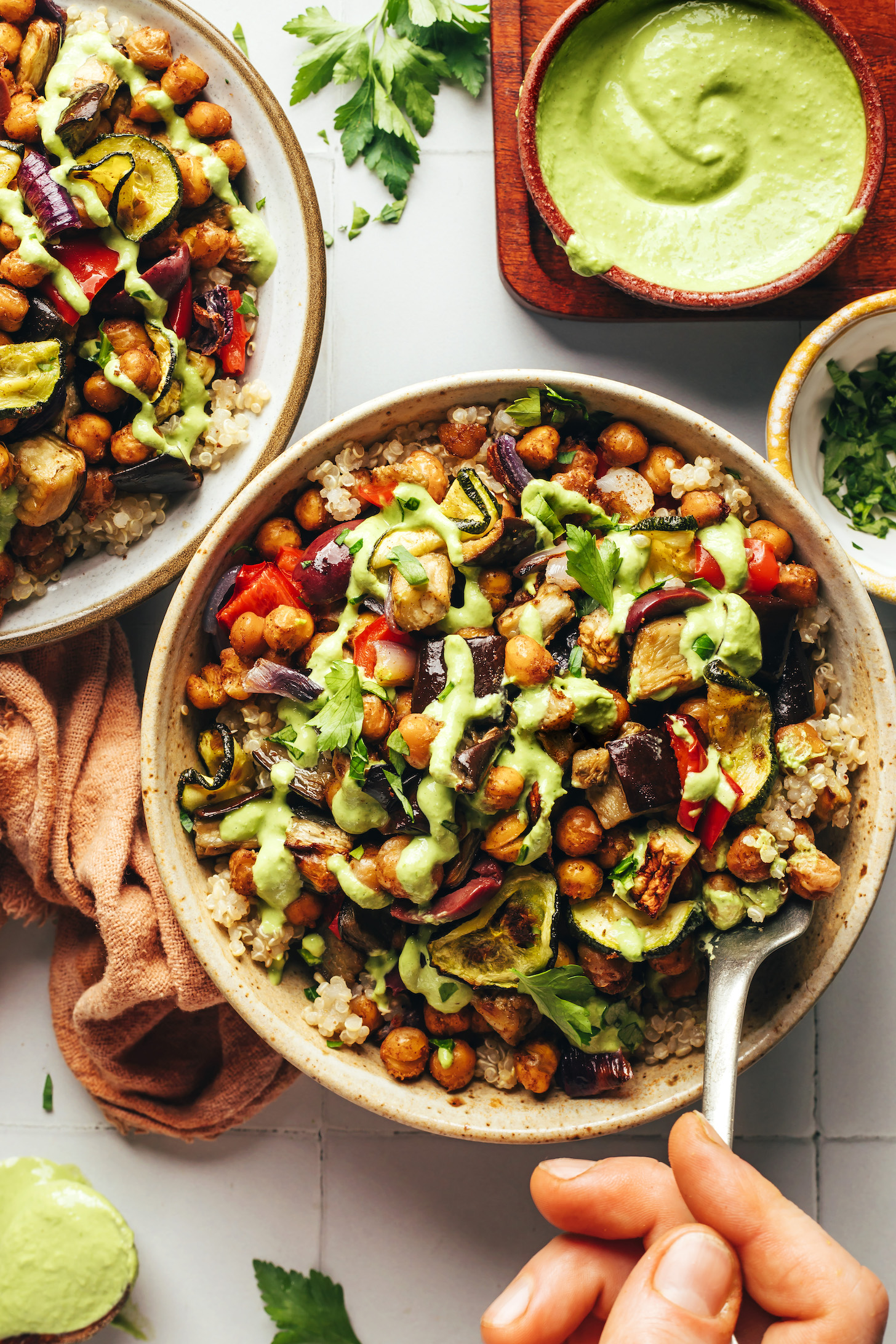 Green tahini sauce drizzled on and in a bowl next to two roasted veggie and chickpea bowls