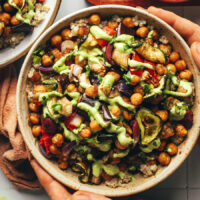 Hands holding a Mediterranean-inspired nourish bowl with chickpeas, veggies, and herby green tahini sauce