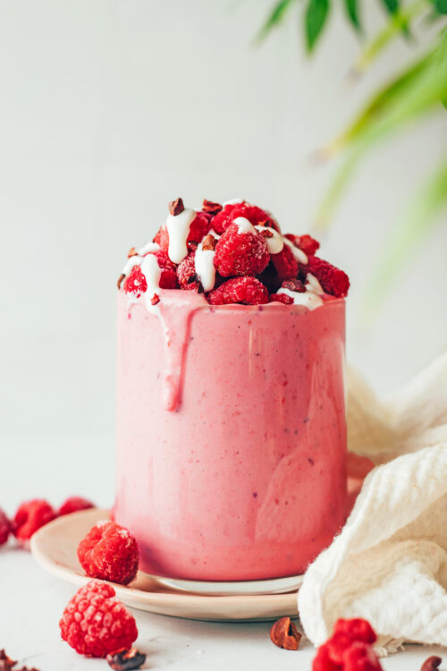Overflowing glass of our raspberry banana smoothie topped with raspberries, coconut yogurt, and cacao nibs