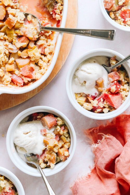 Two bowls of rhubarb crisp topped with vanilla ice cream next to a baking dish with more rhubarb crisp