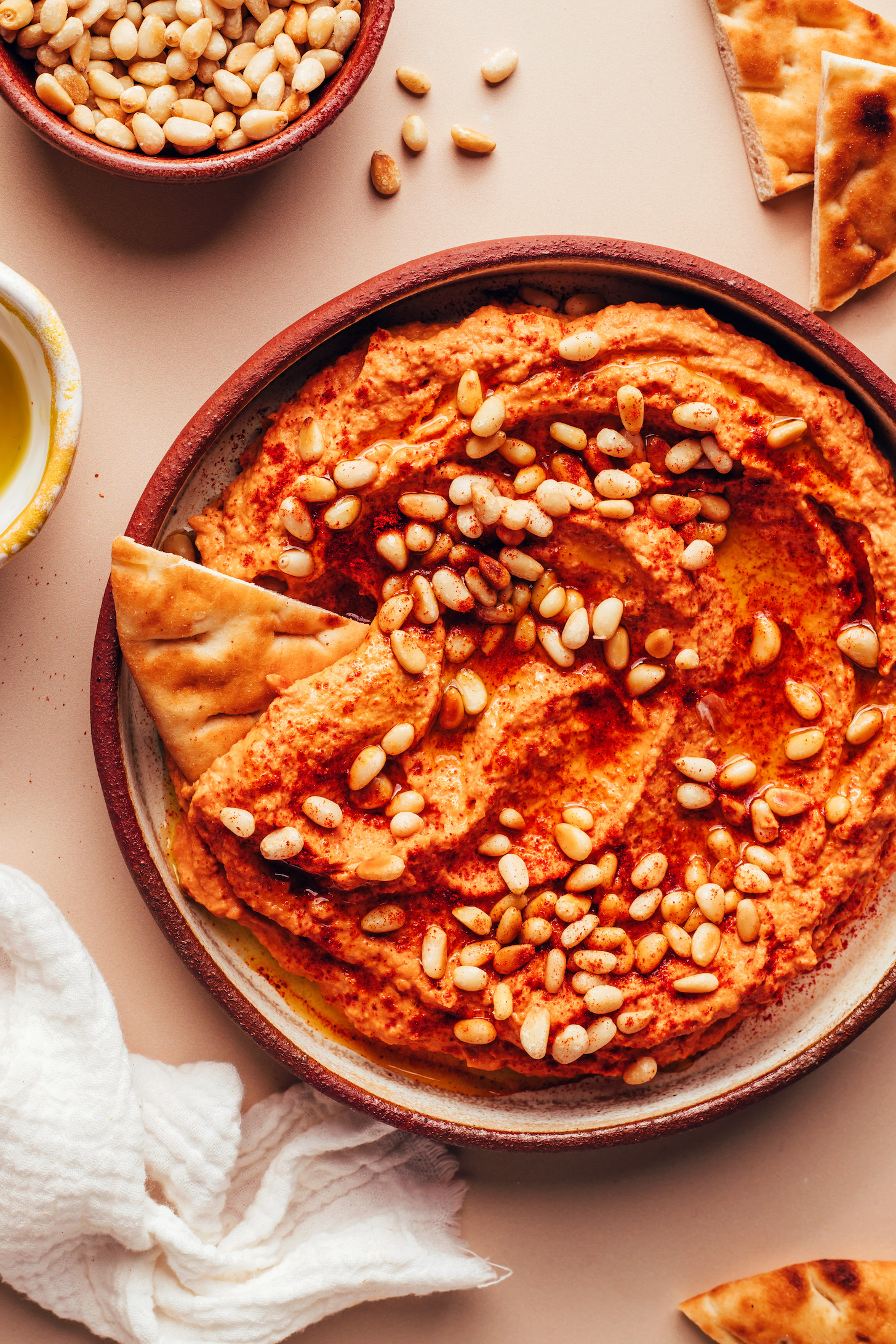 Slice of pita bread dipped into a bowl of sun-dried tomato hummus topped with pine nuts, olive oil, and smoked paprika