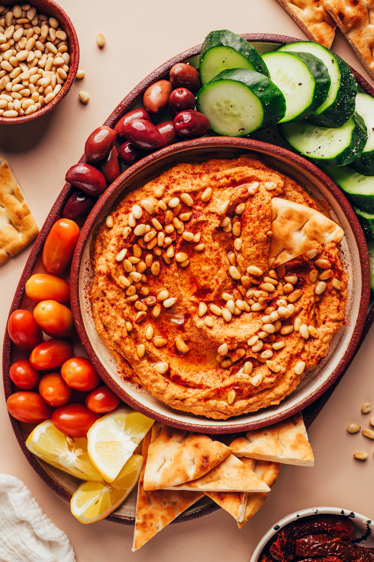 Bowl of sun-dried tomato hummus surrounded by pita bread, lemon slices, baby tomatoes, olives, and cucumber slices