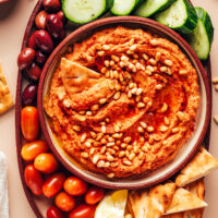 Bowl of sun-dried tomato hummus surrounded by sliced veggies and pita bread for dipping