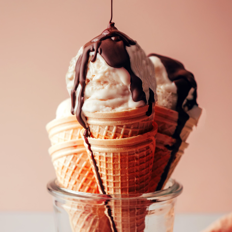 Drizzling chocolate magic shell onto scoops of no-churn vegan vanilla ice cream resting in cones