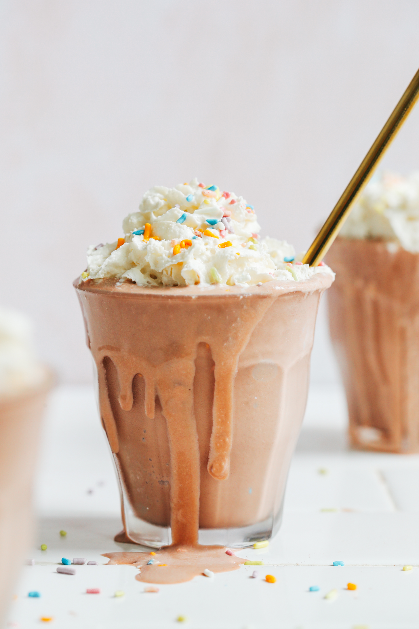 Overflowing glass filled with a vegan chocolate milkshake topped with dairy-free whipped cream and sprinkles