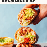 Holding a vegan crunchwrap-inspired burrito above a stack of more burritos