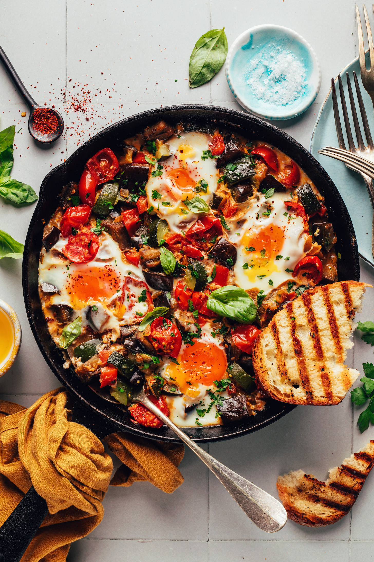 Overhead shot of a slice of bread resting on and beside a pan of skillet ratatouille and eggs