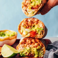 Holding a vegan crunchwrap-inspired burrito above a stack of more burritos