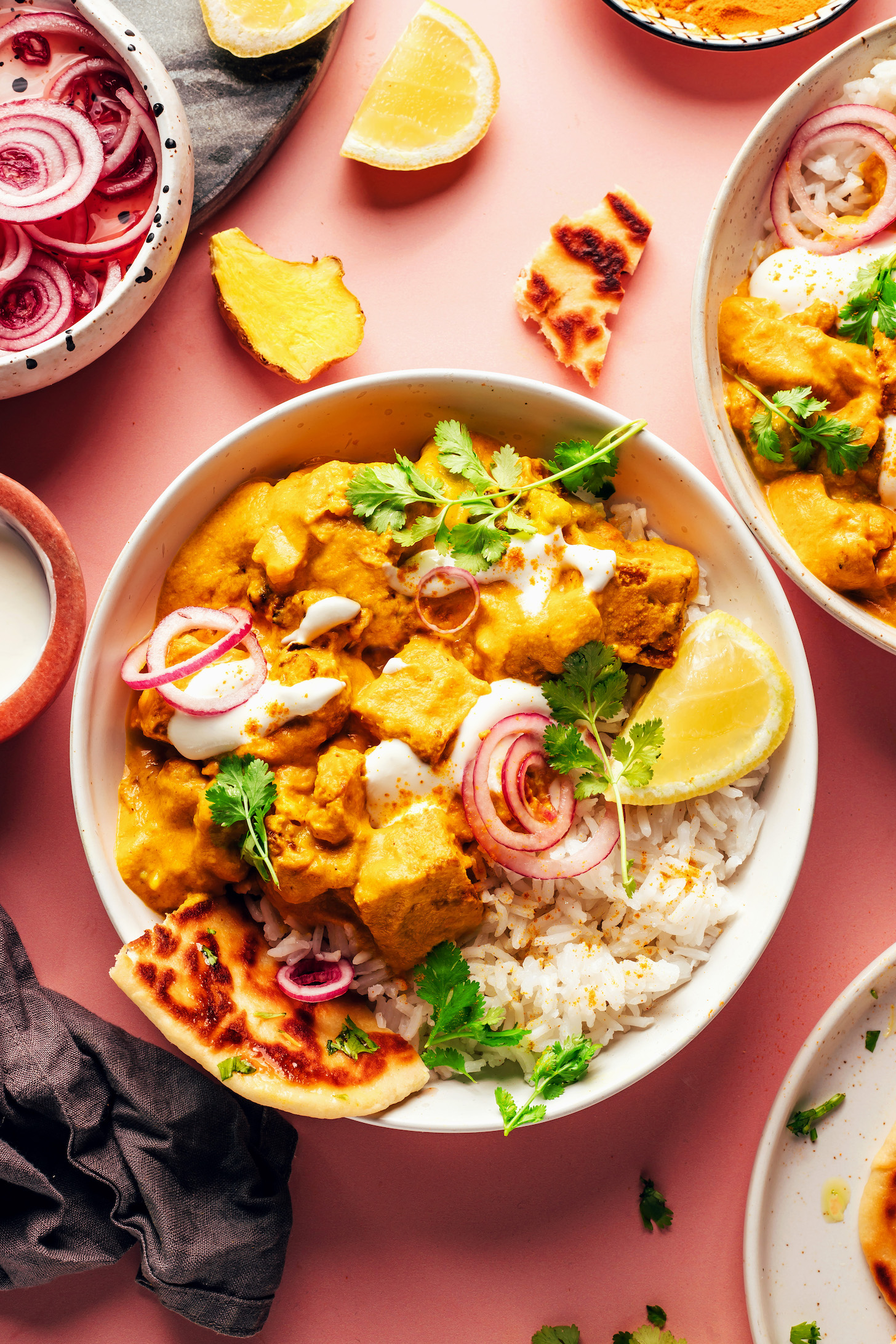 Overhead shot of two bowls of vegan tofu cauliflower korma curry with rice, naan, lemon wedges, pickled onions, and cilantro