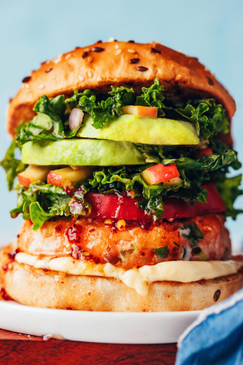 Close up photo of a loaded salmon burger with mayonnaise, tomato, kale apple slaw, and avocado