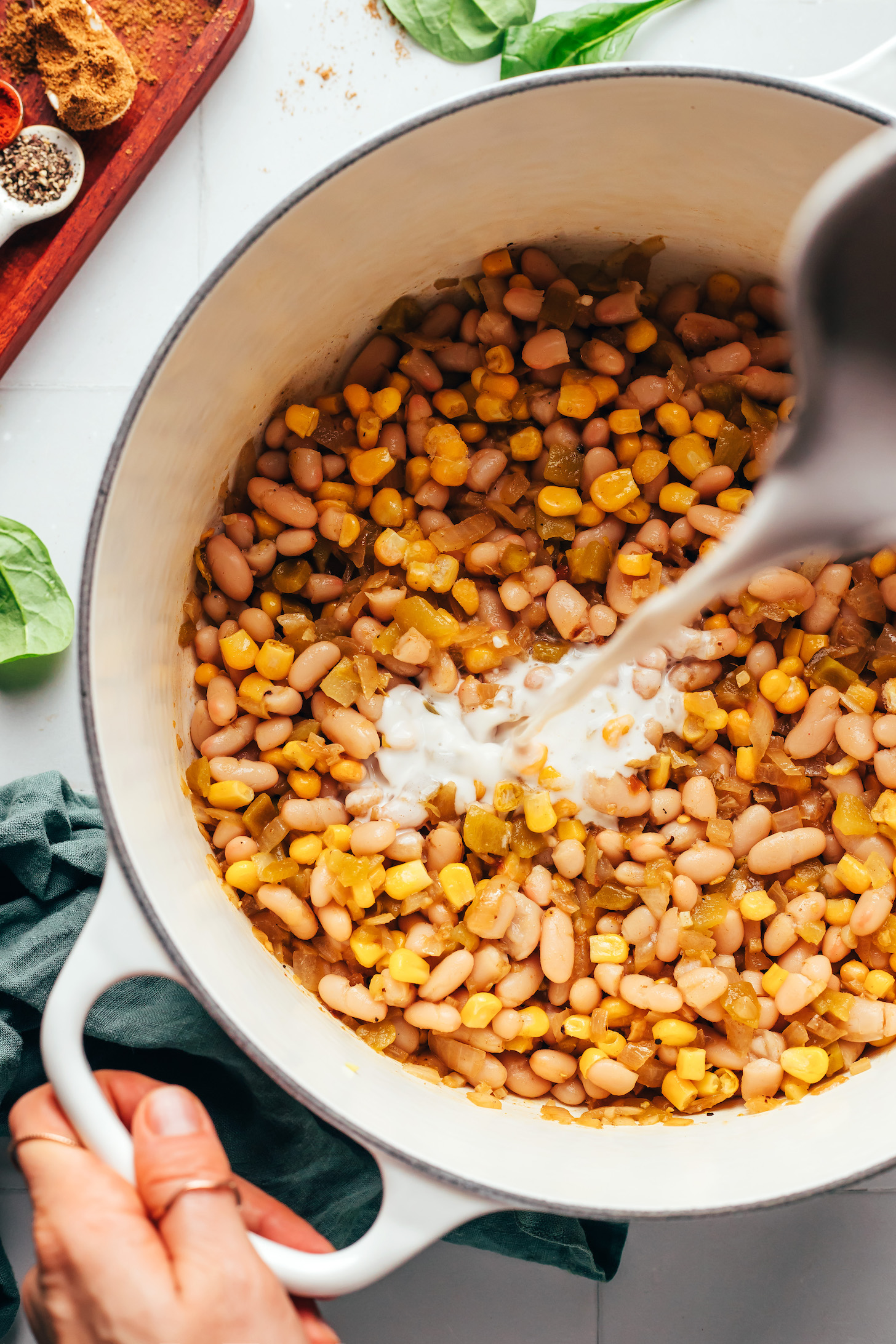 Pouring homemade cashew milk over white beans, green chiles, and corn in a Dutch oven