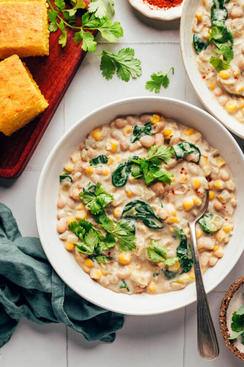 Spoon in a bowl of creamy vegan white bean chili next to cilantro and slices of cornbread