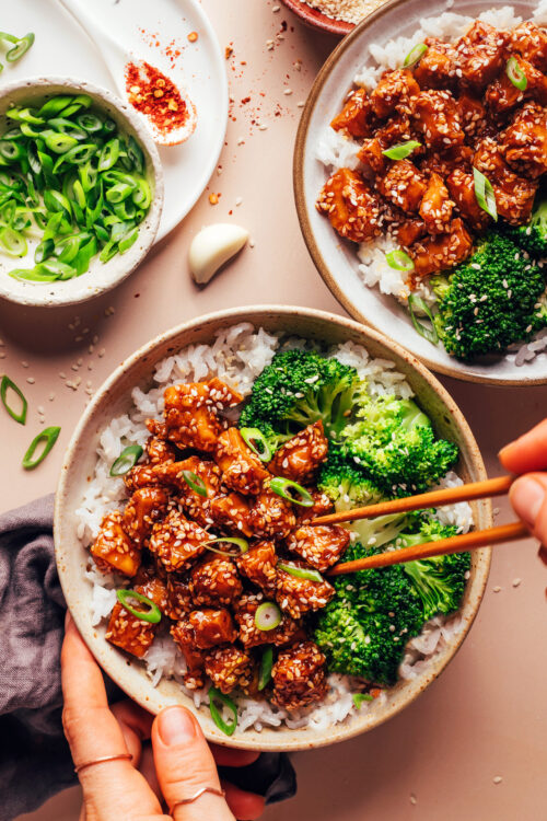 Using chopsticks to pick up a bite of sesame tofu from a bowl with rice and steamed broccoli