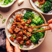 Using chopsticks to pick up spicy garlicky sesame tofu from a bowl of rice and broccoli