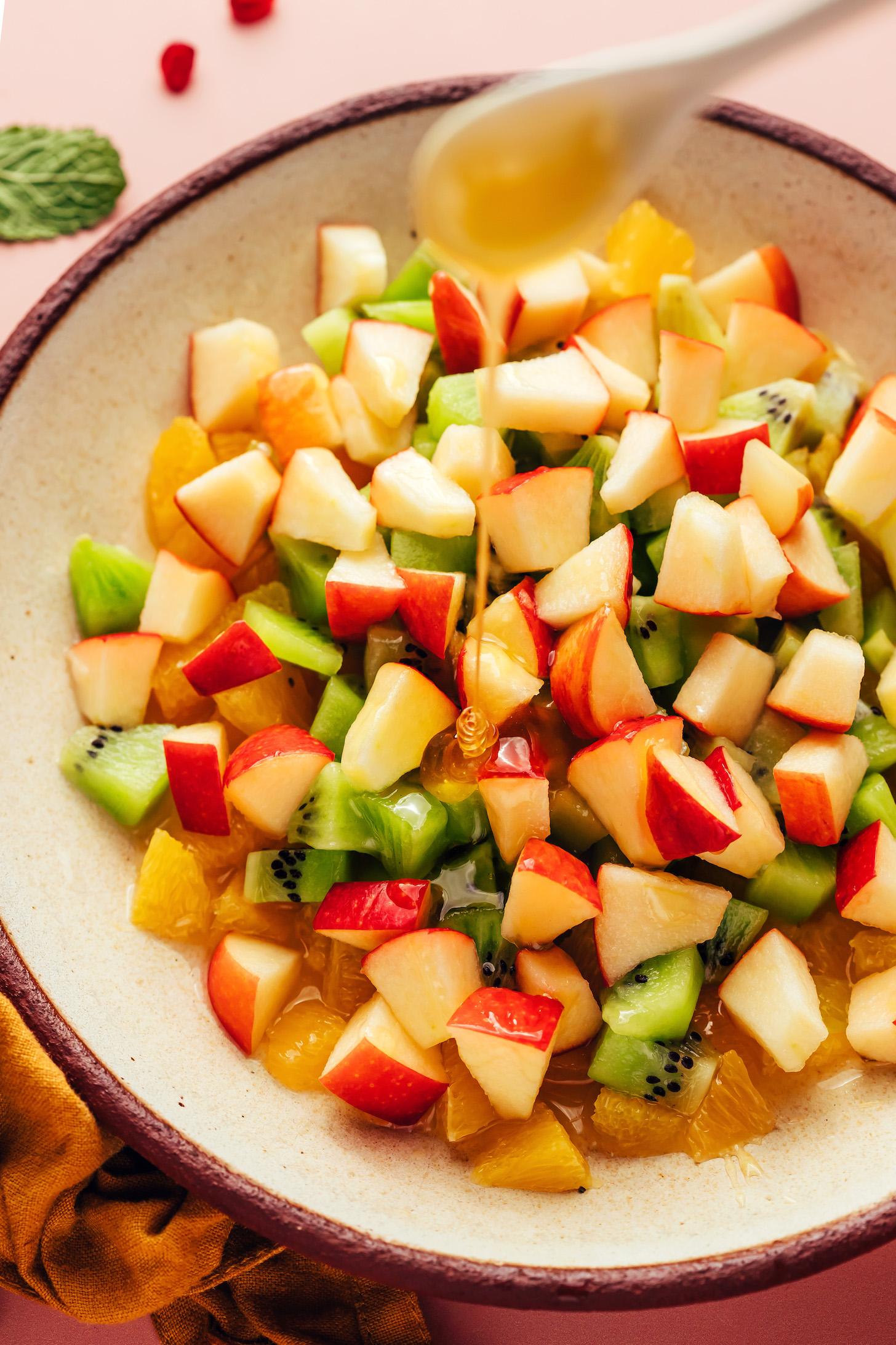 Drizzling honey into a bowl of chopped apple, kiwi, and orange