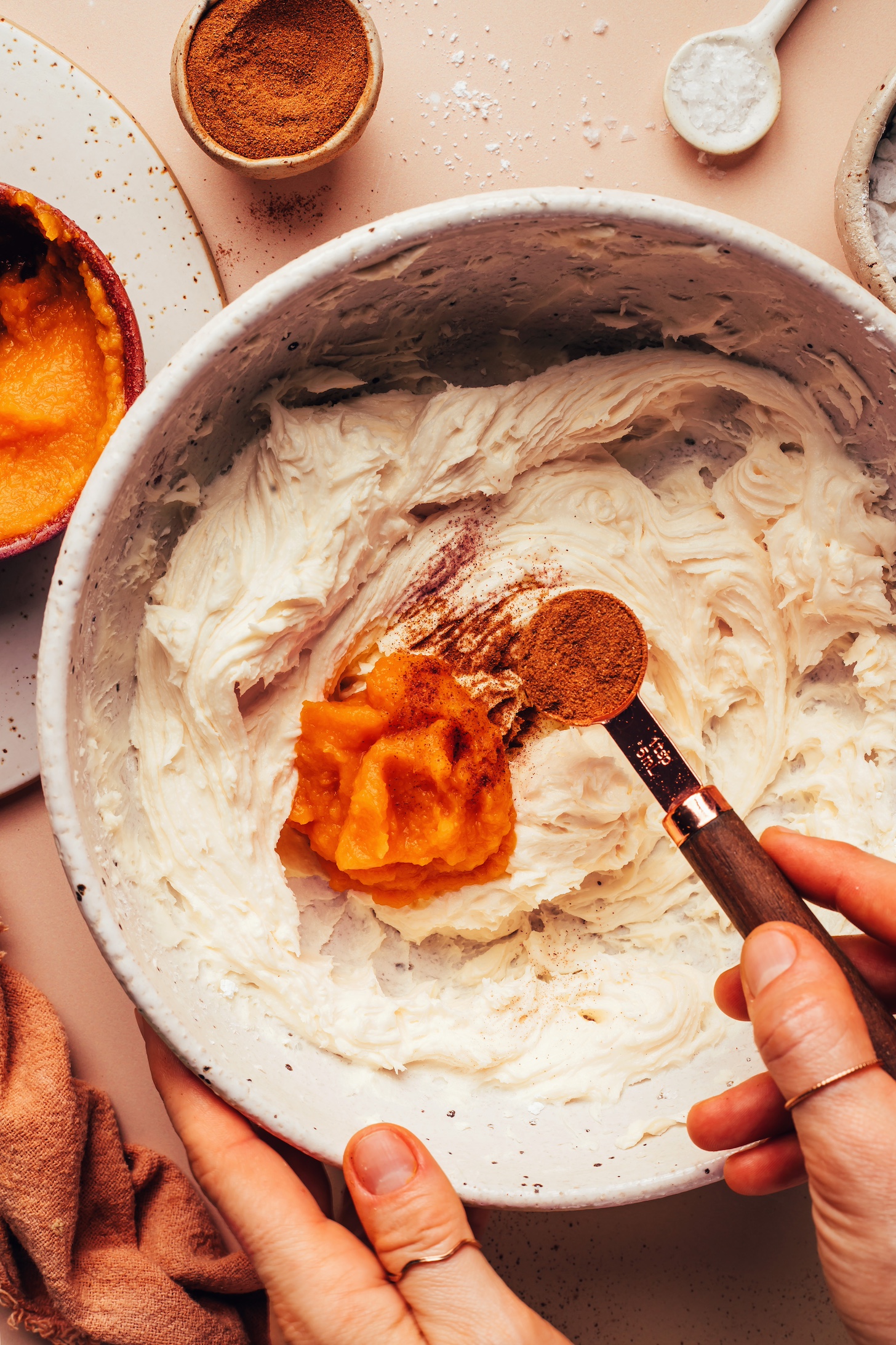 Adding pumpkin pie spice to a bowl of vegan buttercream topped with a scoop of pumpkin purée