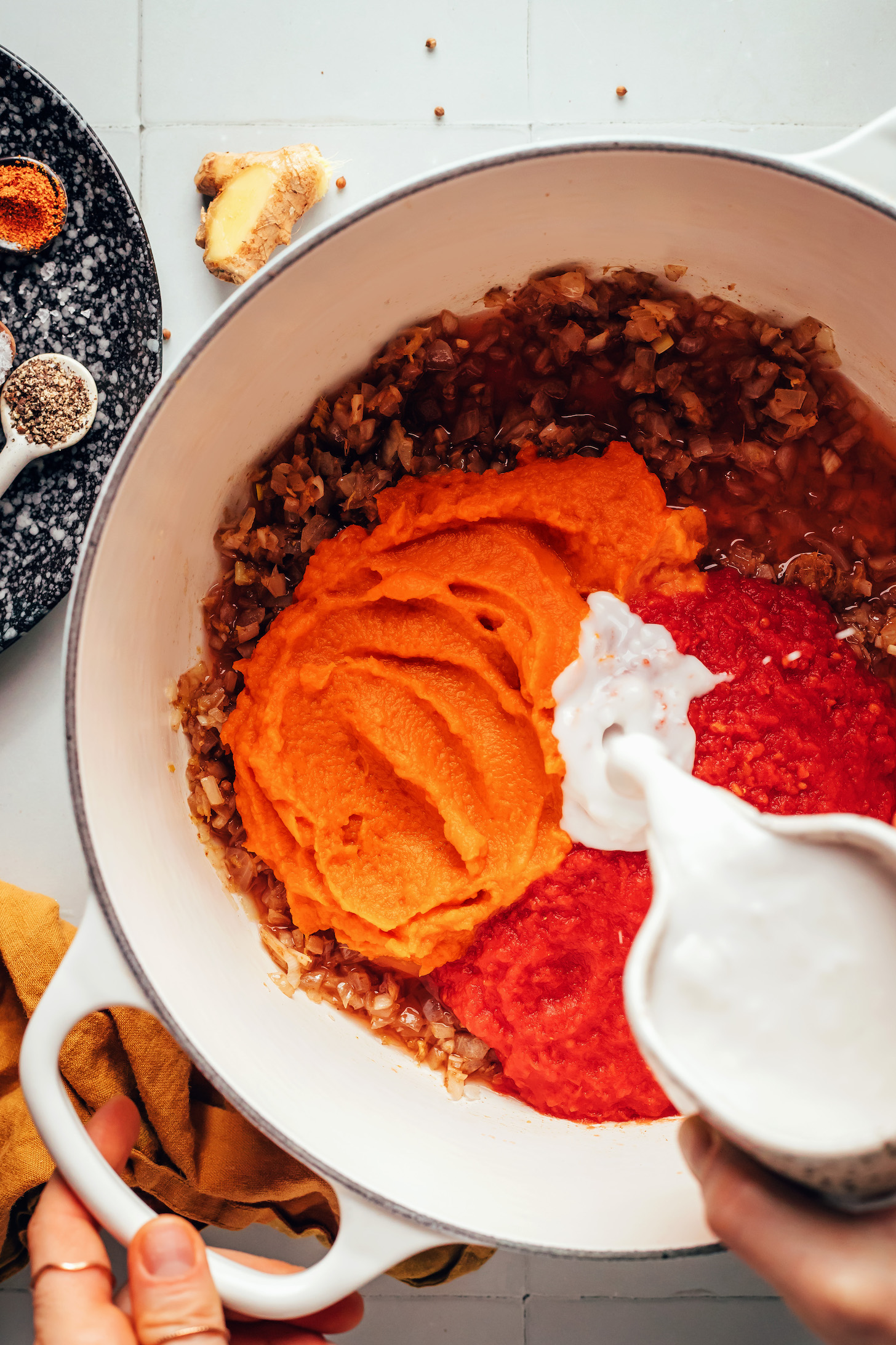 Pouring coconut milk over tomatoes, pumpkin, and sautéed aromatics in a Dutch oven